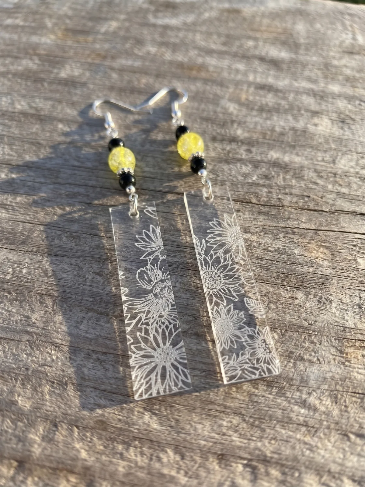 Pair of clear acrylic sunflower drop earrings with yellow and black beads on weathered wood