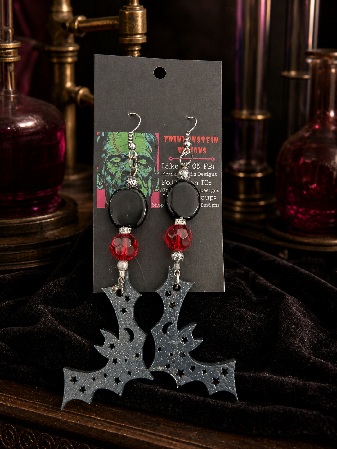 Wooden carved bat earrings displayed on a Frankenstein Designs card in a nocturnal laboratory scene